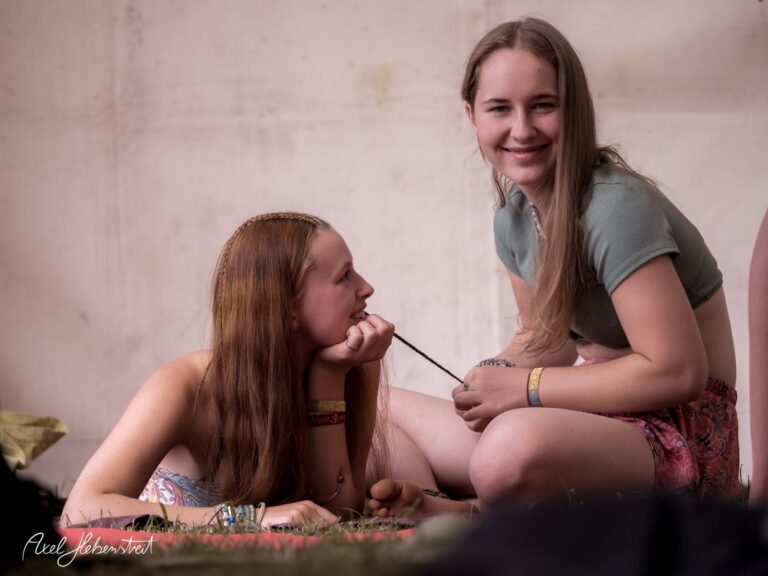 Zwei Mädchen entspannen sich auf einer Wiese, während eines Festivals, eine flechtend, die andere lächelnd. – © Axel Hebenstreit