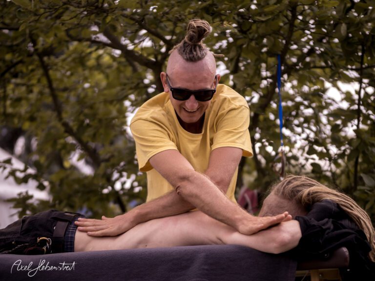 Ein Mann mit einem Dutt massiert entspannt einen anderen am Festival, umgeben von Bäumen. – © Axel Hebenstreit
