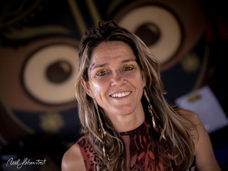 Lächelnde Frau mit Glitzer im Gesicht vor einem bunten Hintergrund, der eine fröhliche Festivalstimmung vermittelt – © Axel Hebenstreit