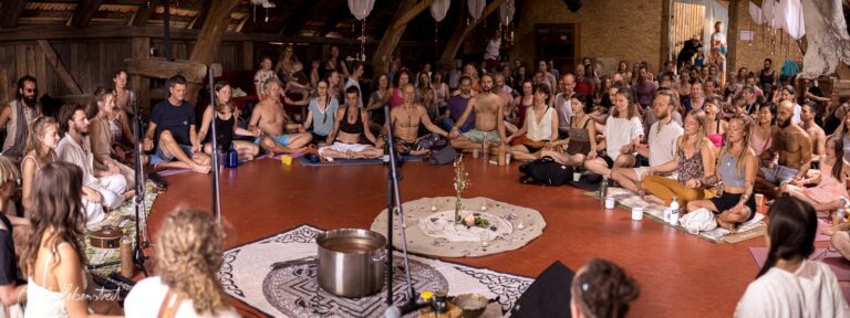 Eine große Gruppe von Menschen sitzt meditativ in einem Saal, umgeben von naturbelassener Holzarchitektur – © Axel Hebenstreit