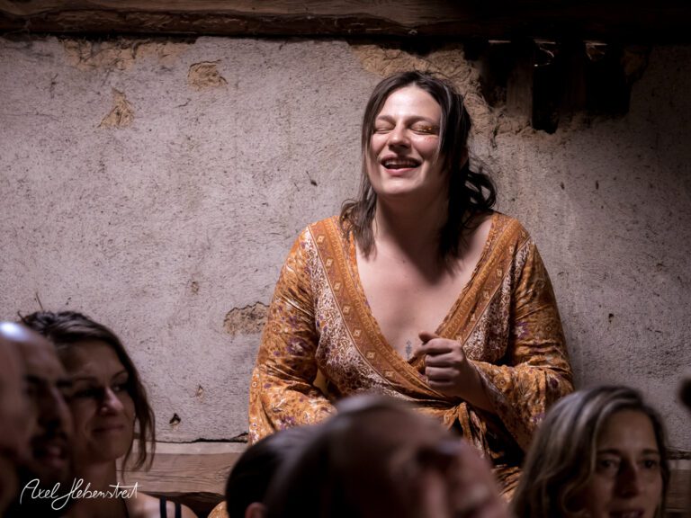 Frau in einem rustikalen Raum, lächelt und genießt den Moment, umgeben von amüsierten Zuschauern – © Axel Hebenstreit