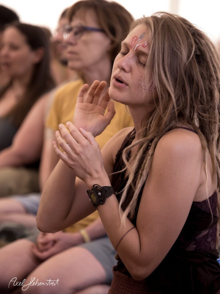 Eine Frau mit Dreadlocks und Facepaint sitzt in meditativer Stimmung bei einem Festival. – © Axel Hebenstreit