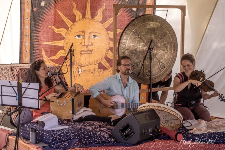 Drei Musiker spielen vor einem bunten Hintergrund mit Sonne, während sie verschiedene Instrumente in einem Zelt nutzen – © Axel Hebenstreit.