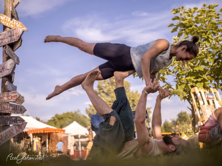 Akrobatische Yoga-Übung im Freien, mit einer Person, die in der Luft gehalten wird, umgeben von festivalähnlicher Atmosphäre – © Axel Hebenstreit