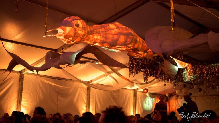 Eine beleuchtete, künstlerische Fischinstallation schwebt über den Gästen in einem festlichen Zelt. – © Axel Hebenstreit