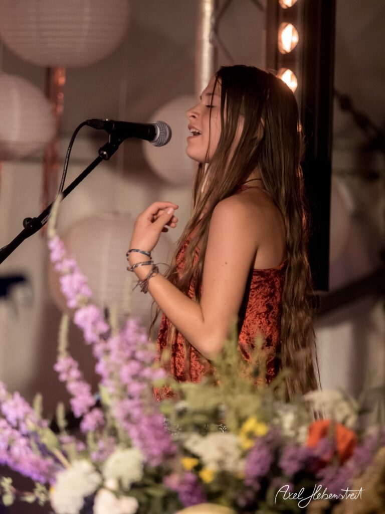 Junge Sängerin mit langen Haaren performt leidenschaftlich auf einer Bühne, umgeben von Blumen – © Axel Hebenstreit