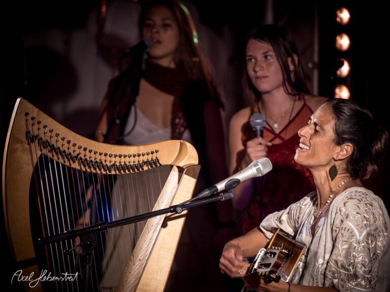 Eine Musikerin spielt Gitarre und singt, während sie von zwei Sängerinnen begleitet wird – © Axel Hebenstreit