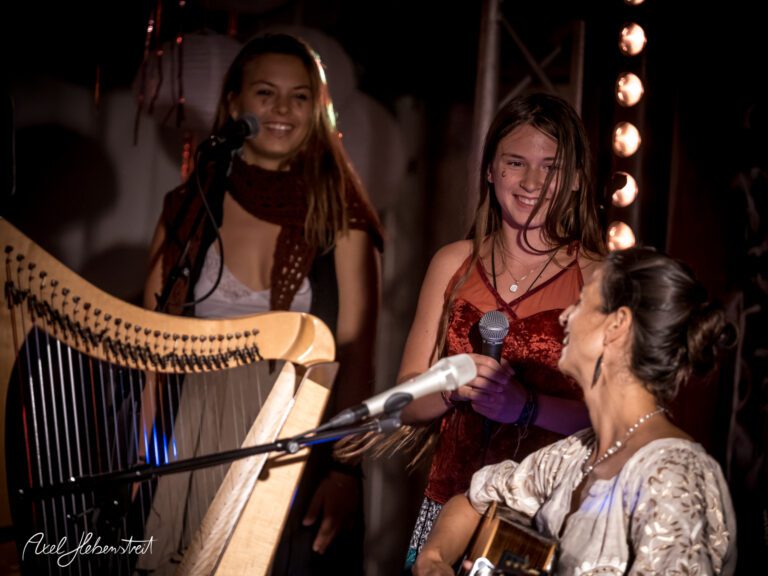 Zwei junge Frauen singen und lächeln fröhlich auf einer Bühne, während eine Musikerin am Harfe spielt – © Axel Hebenstreit