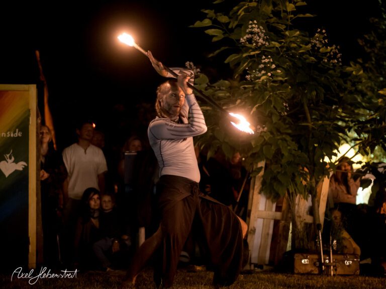 Darbietung eines Feuerakrobaten bei Nacht unter freiem Himmel, umgeben von begeistertem Publikum – © Axel Hebenstreit