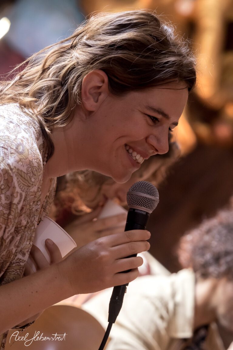 Lächelnde Frau mit Mikrofon, die in eine fröhliche Runde spricht, festgehalten in einer lebendigen Atmosphäre – © Axel Hebenstreit