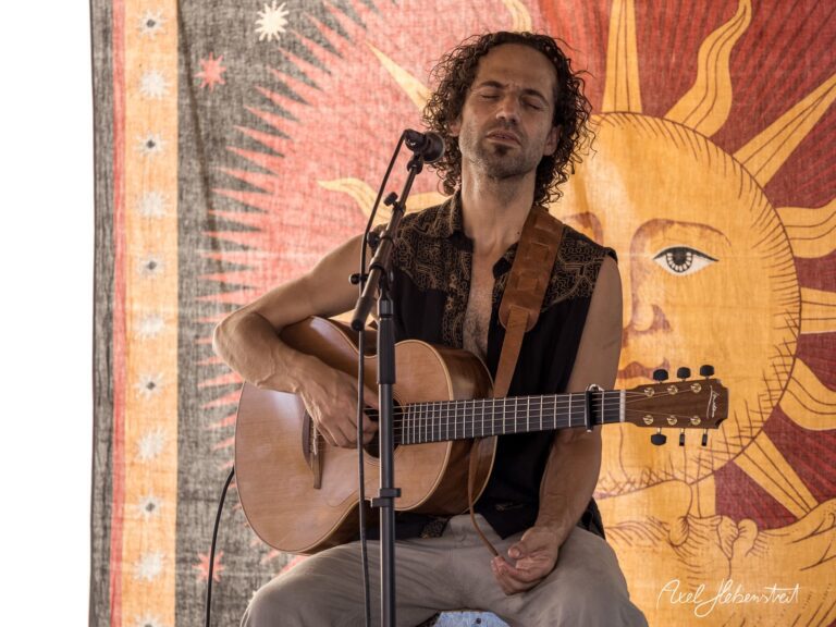 Musiker spielt akustische Gitarre mit geschlossenen Augen, im Hintergrund eine sonnige Wanddekoration – © Axel Hebenstreit