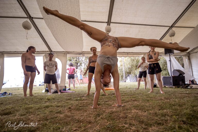 Eine Person führt einen Handstand in einem Zelt auf einem Festival aus, während andere Zuschauer beobachten. – © Axel Hebenstreit