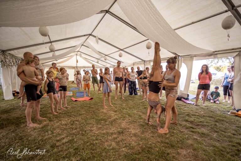 Yoga-Workshop unter einem Zelt, bei dem Teilnehmer akrobatische Übungen vor einer interessierten Gruppe vorführen – © Axel Hebenstreit.