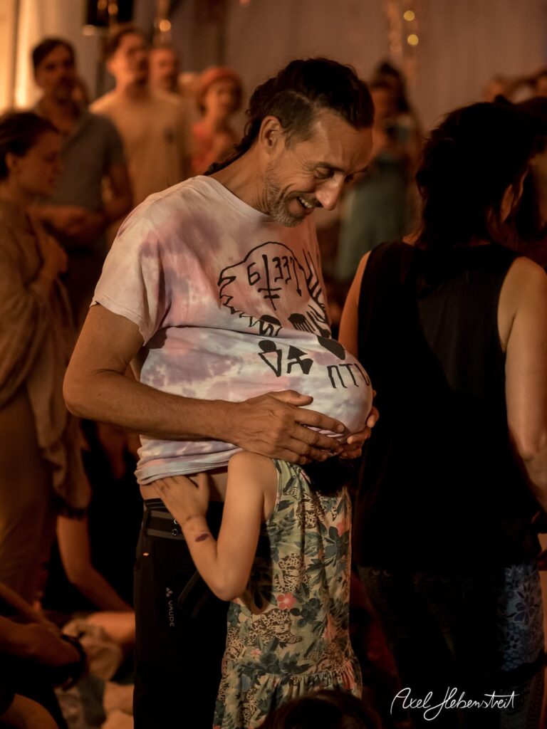 Ein lächelnder Mann umarmt ein Kind, während sie sich inmitten einer geselligen Menge in einem warmen Raum befinden. – © Axel Hebenstreit