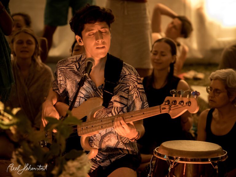 Ein Musiker spielt Gitarre und singt inmitten einer entspannten Festivalatmosphäre. – © Axel Hebenstreit