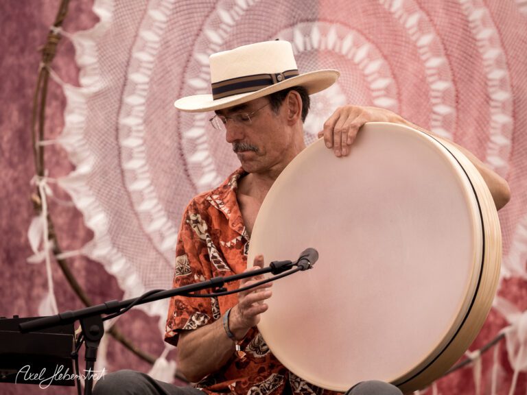 Ein Musiker in einem bunten Hemd spielt einen Rahmen mit einem Mikrofon vor einem bunten Hintergrund – © Axel Hebenstreit