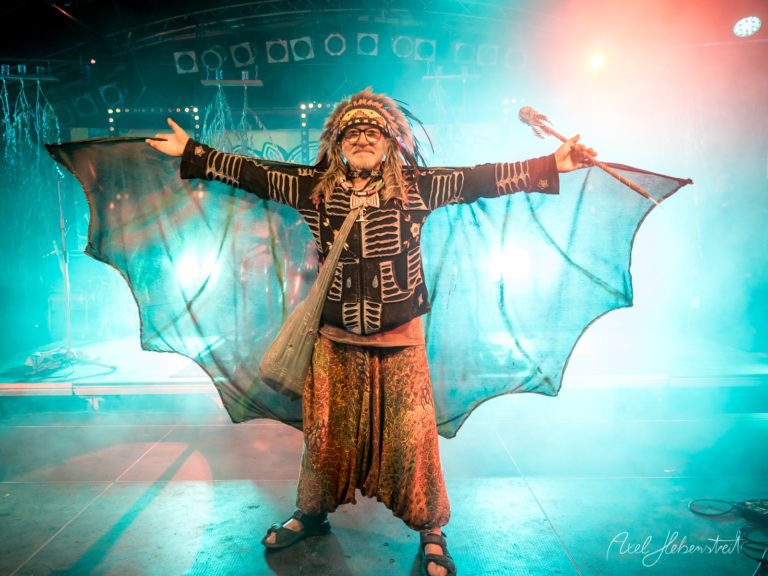 Ein fantasievoller Performer mit Flügeln und Trommel hebt die Arme in einer bunten Festivalatmosphäre – © Axel Hebenstreit