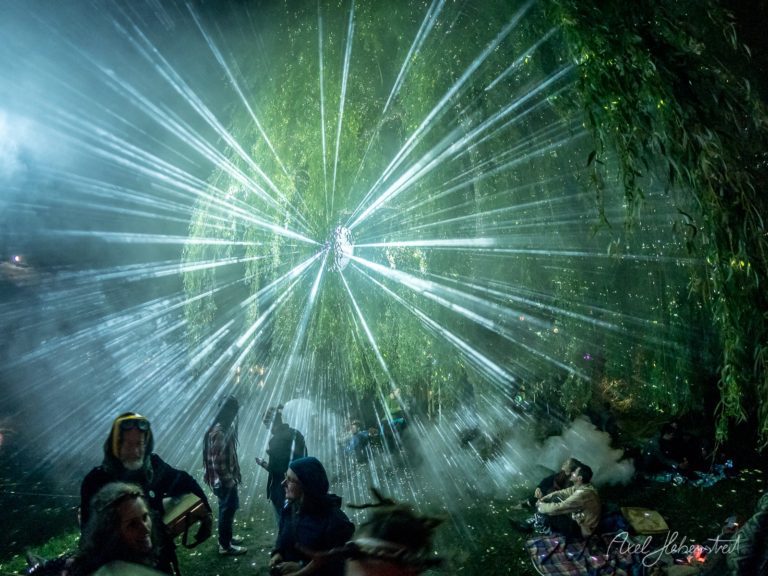 Strahlende Lichtprojektionen durch einen Baum bei einem Festival mit feiernden Menschen – © Axel Hebenstreit