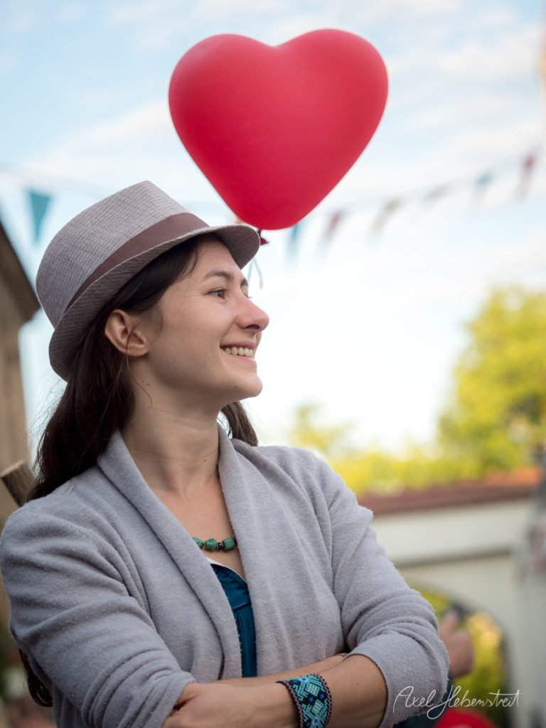 Frau mit Hut und Herzluftballon lächelt verträumt auf einem Festival im Freien – © Axel Hebenstreit