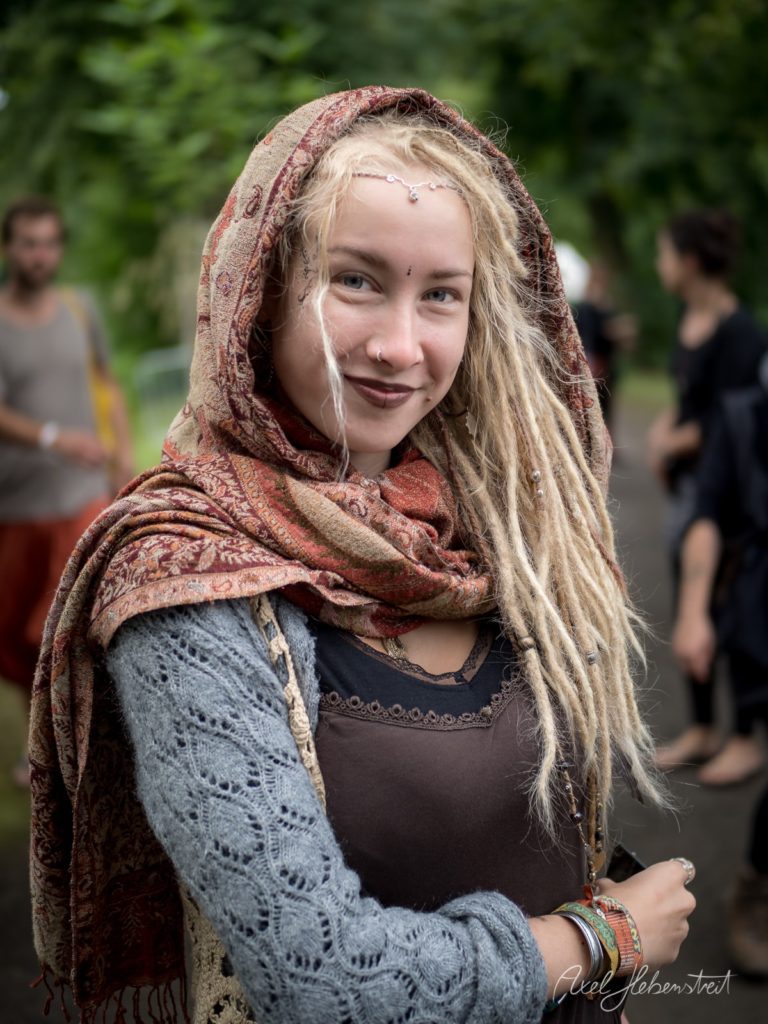 Junge Frau mit Dreadlocks und Tuch, lächelt in einer grünen, lebhaften Festivalatmosphäre – © Axel Hebenstreit