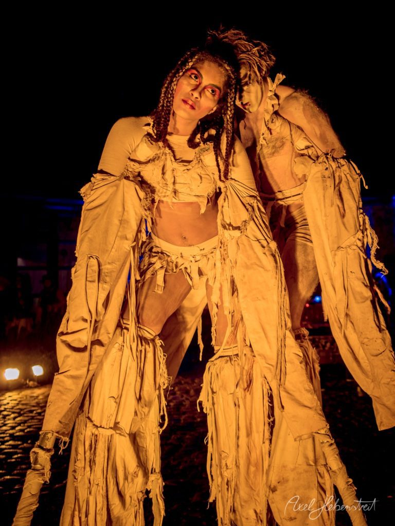Zwei Performer in kreativen Kostümen stehen in einer dramatischen Pose vor dunklem Hintergrund – © Axel Hebenstreit
