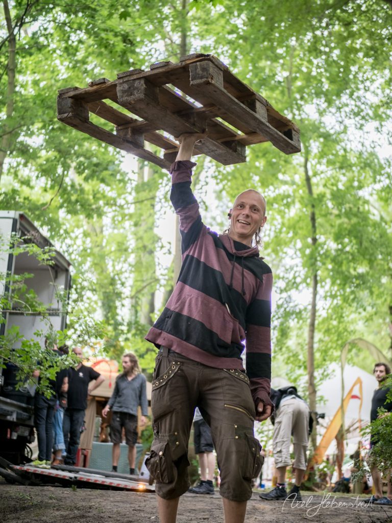 Ein fröhlicher Mann hebt ein Holzpalette in einer grünen, festivalartigen Umgebung. – © Axel Hebenstreit