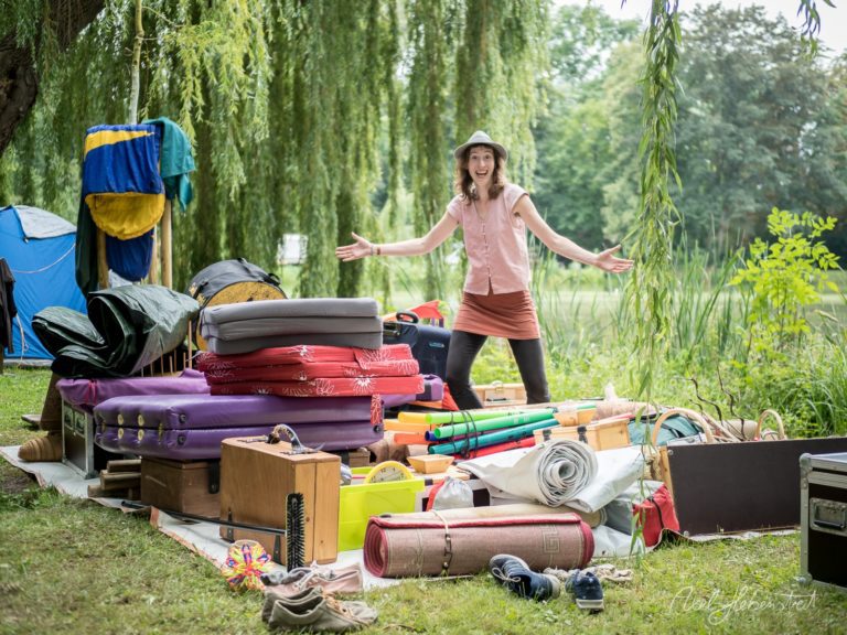 Eine fröhliche Person posiert spielerisch zwischen einem bunten Haufen von Campingutensilien am Wasser – © Axel Hebenstreit.