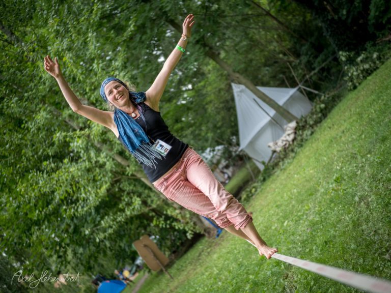Frau balanciert barfuß auf einem Slackline im Grünen, umgeben von Bäumen und einem Festivalambiente – © Axel Hebenstreit