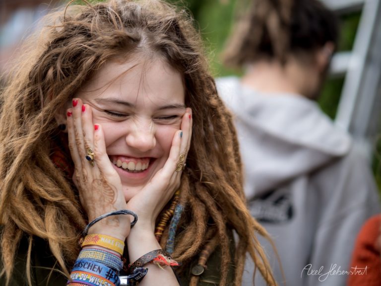 Lächelnde junge Frau mit Dreadlocks hält sich überrascht das Gesicht, umgeben von Festivalatmosphäre – © Axel Hebenstreit