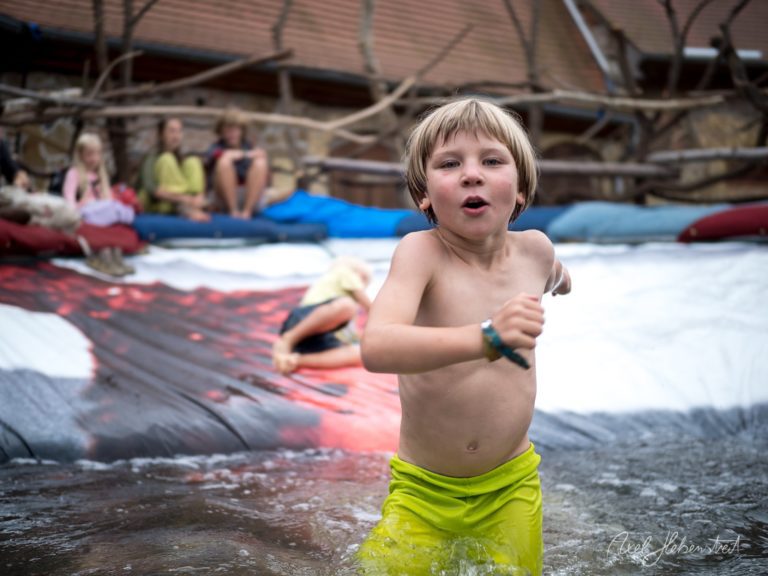 Junge mit blonden Haaren läuft fröhlich in einem Wasserspielbereich, im Hintergrund sitzen entspannte Personen – © Axel Hebenstreit