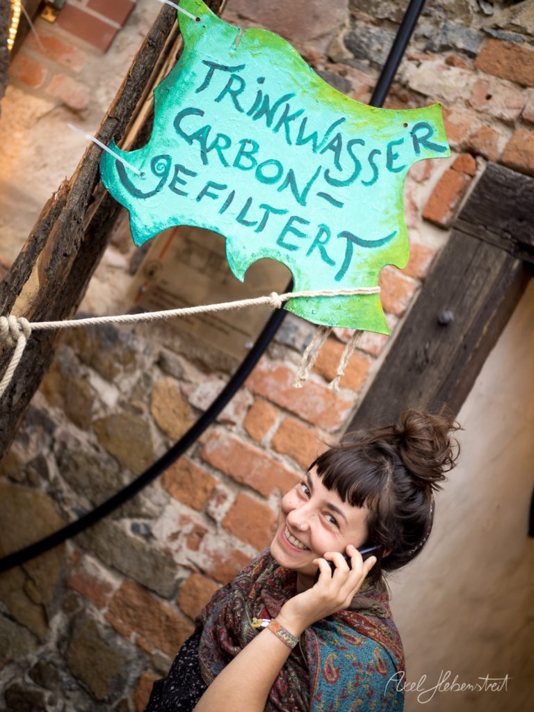 Lächelnde Frau, die mit dem Handy telefoniert, neben einem handgemalten Schild für gefiltertes Trinkwasser – © Axel Hebenstreit