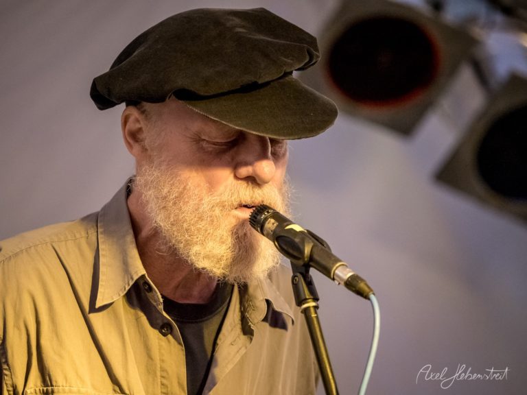 Ein Mann mit Bart und Mütze singt in ein Mikrofon, umgeben von Scheinwerfern auf einer Konzertbühne. – © Axel Hebenstreit
