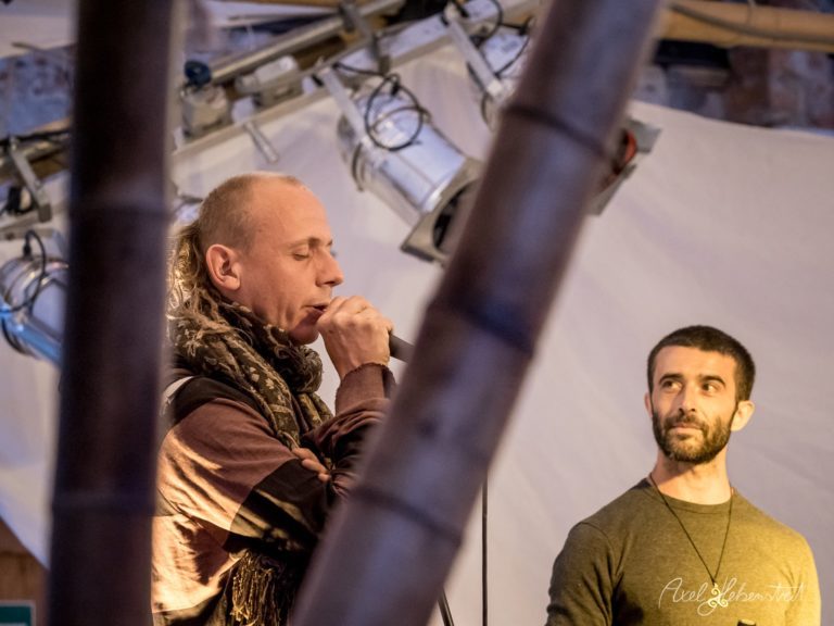 Zwei Musiker auf einer Bühne, einer singt leidenschaftlich ins Mikrofon, während der andere aufmerksam lauscht – © Axel Hebenstreit