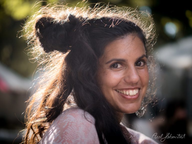 Lächelnde Frau mit zerzausten Haaren, Sonnenlicht und fröhlicher Stimmung im Hintergrund eines Festivals – © Axel Hebenstreit.