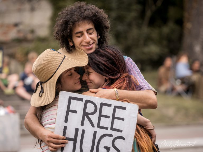 Drei Menschen umarmen sich herzlich und halten ein Schild mit der Aufschrift „FREE HUGS“ – © Axel Hebenstreit