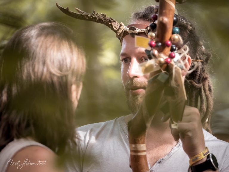 Ein Mann mit Hirschgeweih und Dreadlocks spricht lächelnd mit einer Frau im Wald – © Axel Hebenstreit