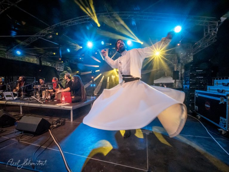 Ein tanzender Derwisch in voller Bewegung auf einer Festivalbühne, umgeben von MusikerInnen – © Axel Hebenstreit