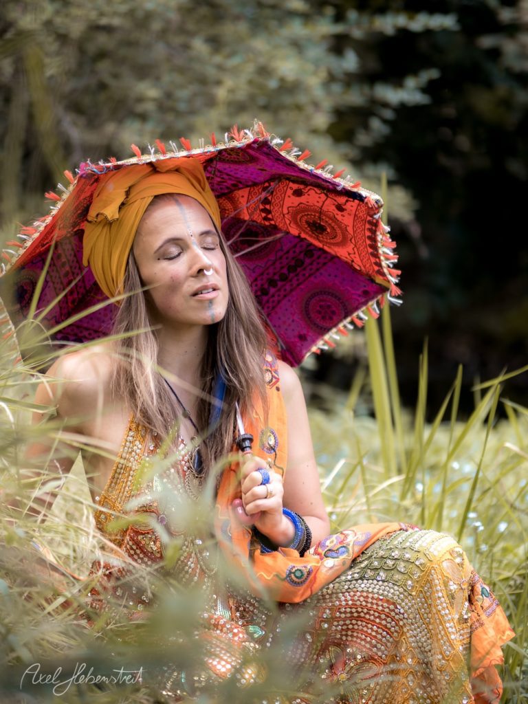 Eine spirituelle Frau in bunten Gewändern meditiert unter einem farbenfrohen Schirm in der Natur. – © Axel Hebenstreit