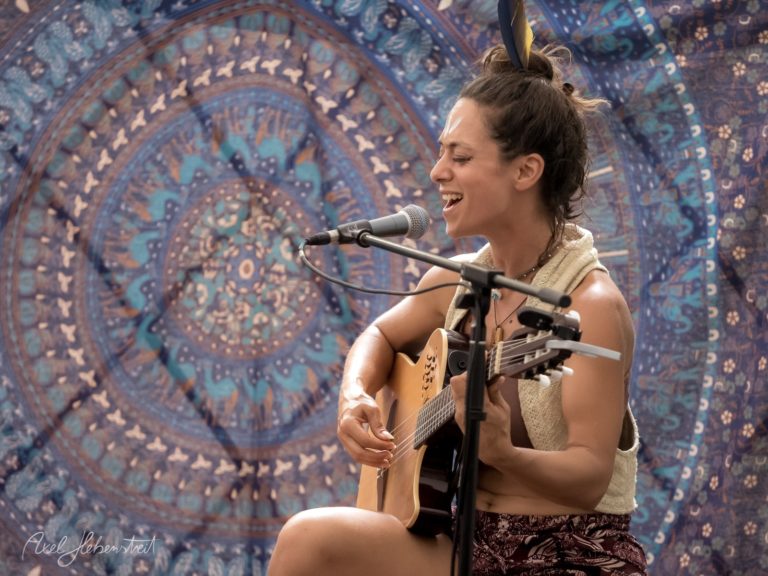 Frau mit Gitarre singt leidenschaftlich vor einem bunten Wandteppich auf einem Festival. – © Axel Hebenstreit