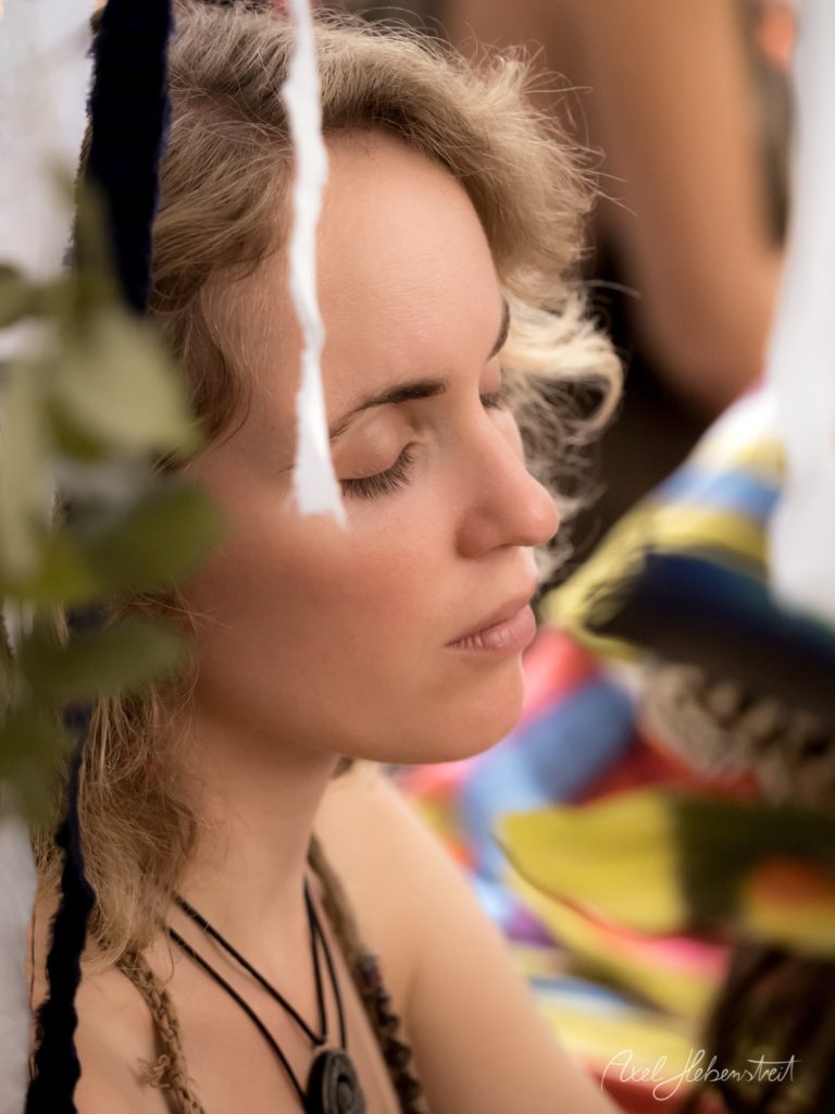 Eine nachdenkliche Frau mit geschlossenen Augen, umgeben von bunten Stoffen und Pflanzen, strahlt Ruhe aus – © Axel Hebenstreit