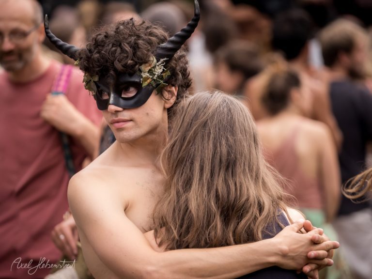 Junger Mann mit Maske und Hörnern umarmt eine Frau auf einem lebhaften Festival in Bayern – © Axel Hebenstreit