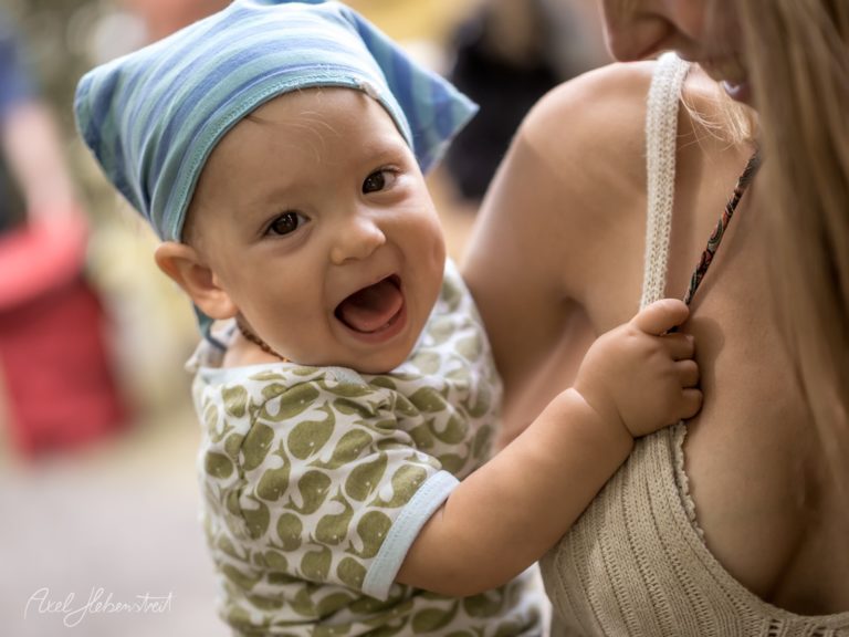 Lachendes Baby in bunter Kleidung und Kopfbedeckung, in den Armen einer Frau, fröhliche Stimmung – © Axel Hebenstreit