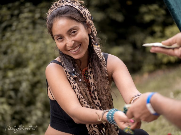 Lächelnde Frau mit Dreadlocks und Tuch, die einem Besucher auf einem Festival etwas reicht – © Axel Hebenstreit
