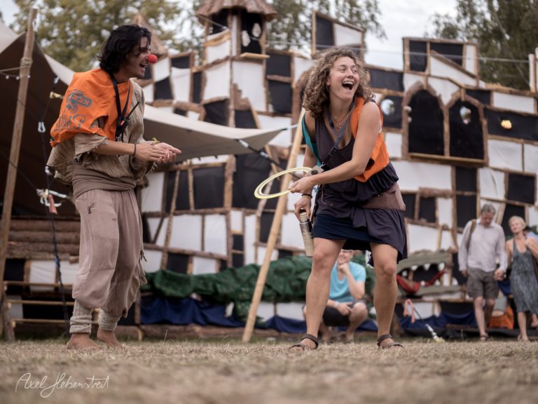 Lachende Menschen in orangefarbenen Westen spielen mit einem Hula-Hoop auf einem Festivalgelände. – © Axel Hebenstreit
