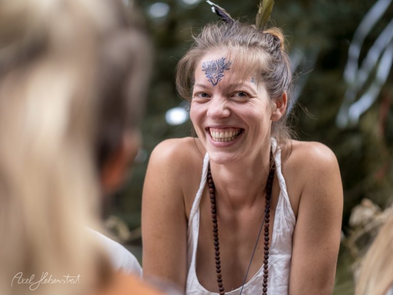 Eine lächelnde Frau mit einem Gesichtstattoo sitzt in einem entspannten Kreis von Freunden in der Natur – © Axel Hebenstreit