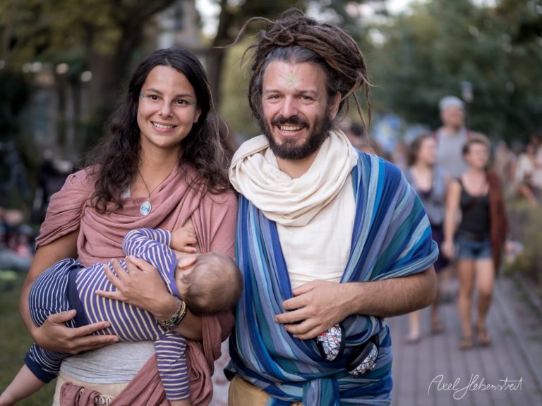 Junges Paar in entspannter Atmosphäre, das ein Baby stillt, während sie in einer Festivalumgebung spazieren. – © Axel Hebenstreit