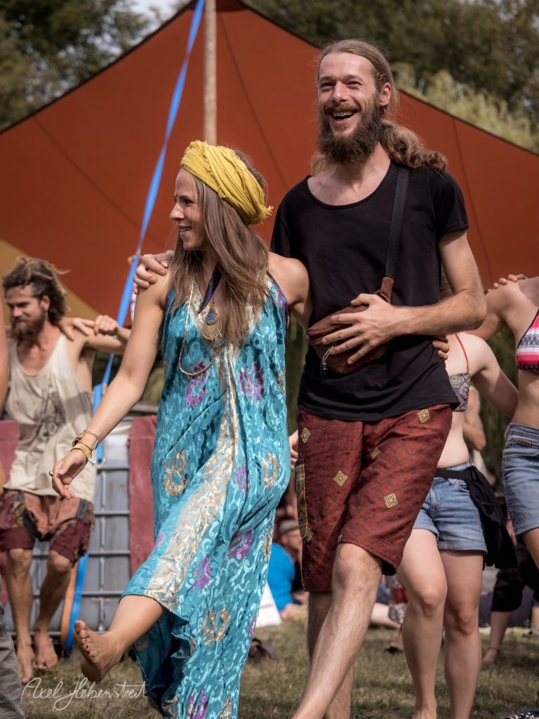 Lächelnd tanzen ein Paar in bunten, festlichen Kleidern auf einem Festival im Freien. – © Axel Hebenstreit