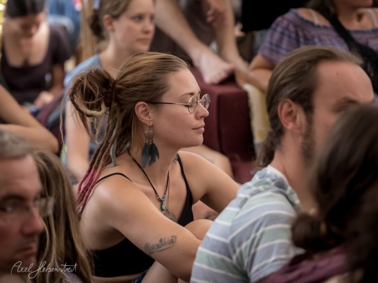 Eine entspannte Frau mit Dreadlocks meditiert inmitten einer großen Gruppe bei einem Fest. – © Axel Hebenstreit
