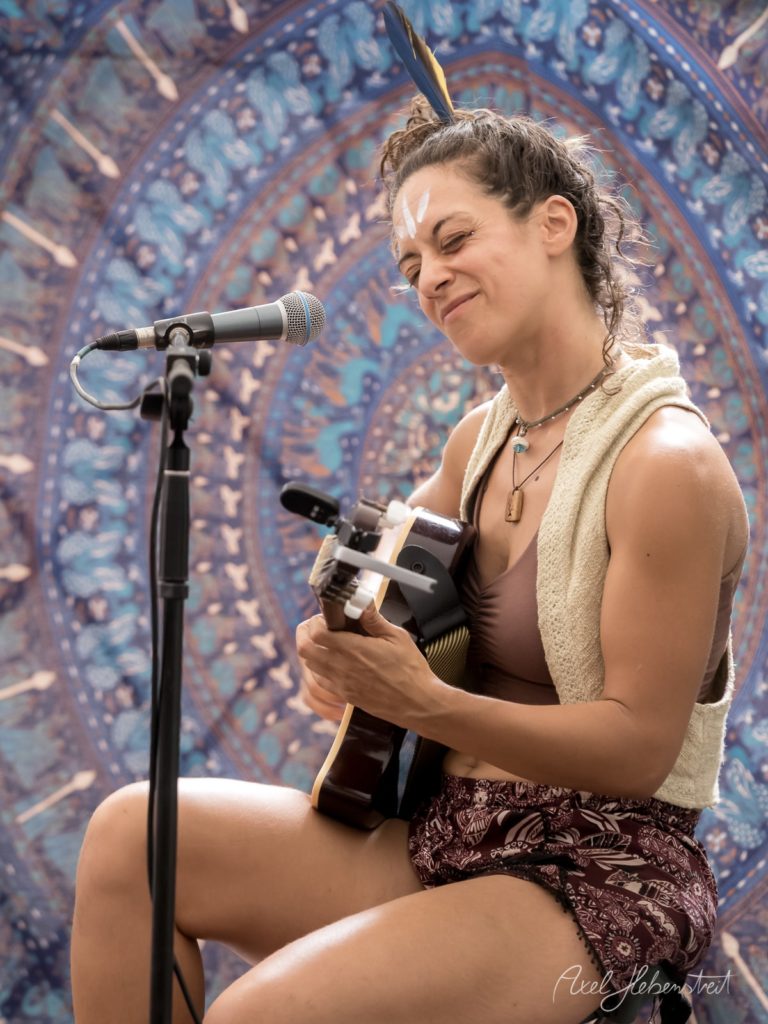 Eine Musikantin mit Gitarre und Feder im Haar spielt leidenschaftlich auf einem Festival. – © Axel Hebenstreit