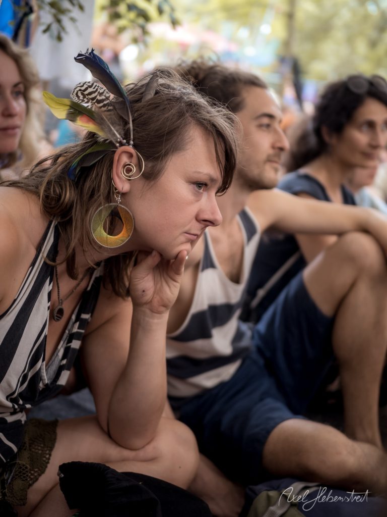 Nachdenkliche Frau mit auffälligen Ohrringen, umgeben von Menschen in einem Festivalambiente – © Axel Hebenstreit